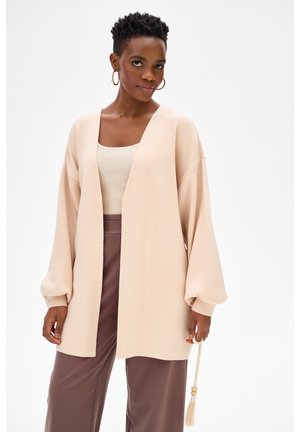 Kobieta z krótkimi kręconymi włosami, ubrana w beżowy otwarty kardigan, kremowy top na ramiączkach, brązowe spodnie o szerokich nogawkach oraz kolczyki koła, trzymająca ozdobę z frędzlami.