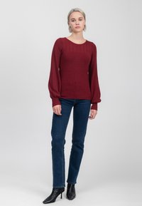Bordeaux gebreid trui met een gestructureerde schouderpartij, lange pofmouwen en een aansluitende silhoeët, gecombineerd met donkerdenim jeans en zwarte geklede laarzen.