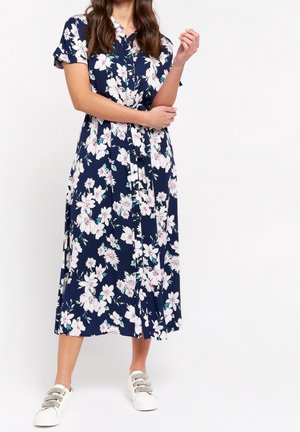 Kobieta w granatowej sukience midi w białe kwiaty, z krótkimi rękawami, zapinana na guziki na przodzie, w białych trampkach, stoi na białym tle.