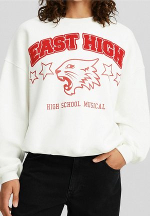 Weißes Sweatshirt mit rotem Schriftzug "East High", einem brüllenden Tigerkopf, Sternen und dem Text "High School Musical", getragen von einer Person mit einer Hand in der Tasche.