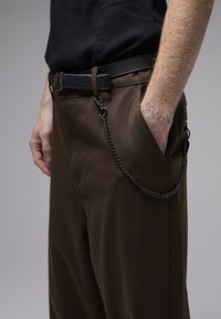 Pantalon marron avec ceinture, doté d'une chaîne en métal attachée à la poche. Design simple et texture lisse, avec des détails de couture visibles.