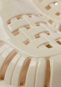 Kreminių spalvų plastikiniai šlepetės su pintu dizainu, turinčiu atvirus plotus ir lygią tekstūrą, užtikrinančią ventiliaciją ir lankstumą formai.