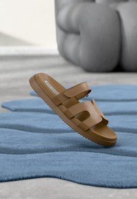 Sandale à glissière en caoutchouc marron avec une semelle épaisse, deux larges brides et un motif découpé sur la partie supérieure, posée sur un tapis bleu texturé.