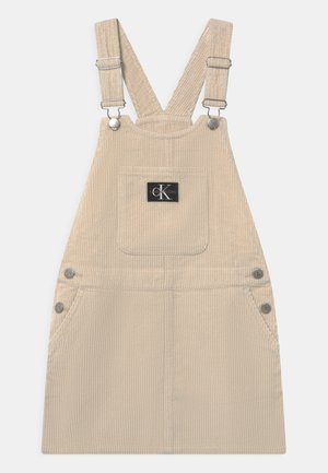 Vestito salopette in velluto a coste beige con bretelle regolabili, tasca frontale e dettagli in metallo argentato. Texture a coste verticali e forma aderente.