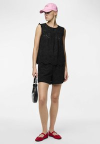 Top noir à œillets, shorts assortis, ballerines rouges et casquette rose. Présente des bords festonnés et une texture réfléchissante sur le sac noir tenu à la main.