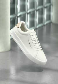 Λευκό δερμάτινο αθλητικό παπούτσι με κομψό σχεδιασμό, στρογγυλή μύτη, υφή στη σόλα και ταμπά διακοσμητικό στη φτέρνα. Κορδόνια και λογότυπο της μάρκας ορατά στη πλευρά.