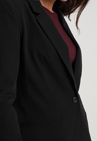 Black tailored blazer with a single button closure, notch lapels, and a slim fit. Features a side pocket and smooth fabric texture.