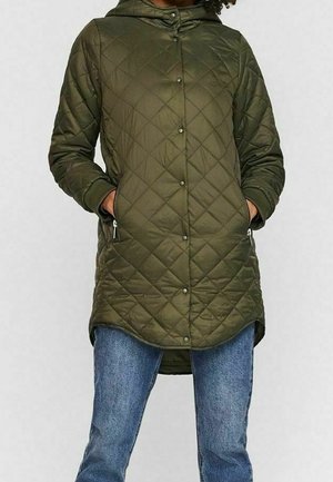 Femme portant une veste matelassée vert olive à capuche avec boutons-pression et poches avant zippées, associée à un jean bleu.