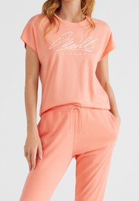 Lekka, koraloworóżowa koszulka z krótkim rękawem i luźnym krojem, z białym napisem logo. W zestawie spodnie z troczkiem w tym samym kolorze.