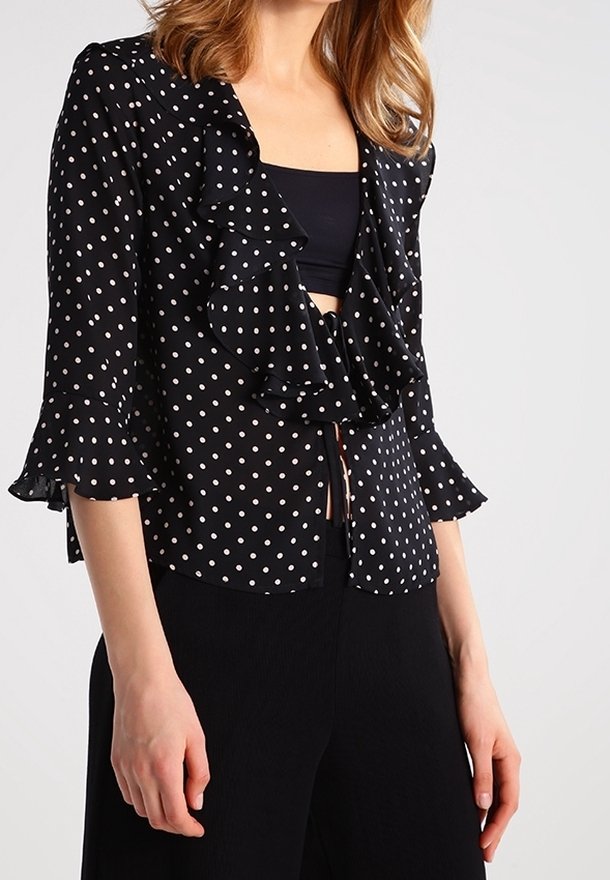 Femme portant un chemisier noir à pois blancs, avec un devant froncé et des manches trois-quarts, par-dessus un top noir, associé à un pantalon noir.