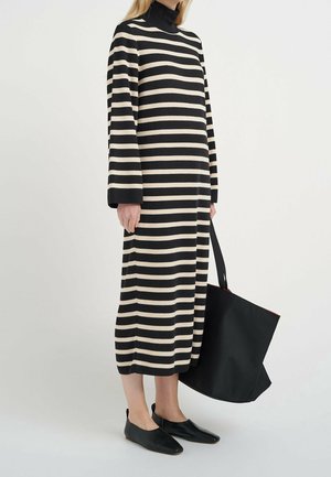 Personne portant une longue robe à rayures horizontales noires et beiges avec des ballerines noires, tenant un grand sac fourre-tout noir à ses côtés.