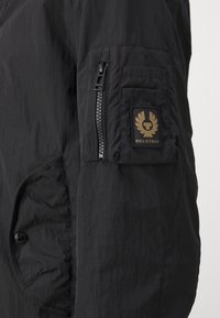 Giacca nera realizzata in tessuto leggero, con una tasca con zip sulla manica e un distintivo logo dorato con un design di aquila caratteristico.