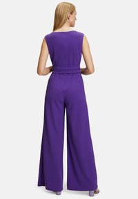 Lila ujjatlan overall széles lábakkal és övvel a deréknál. Sima anyag, egyszerű dizájn, hátul gombos záródással.