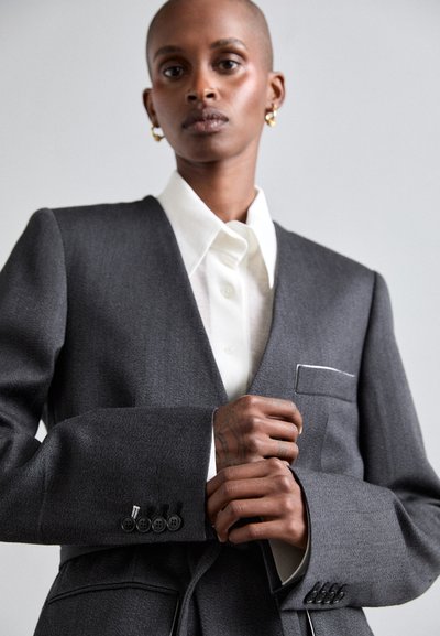 Szürke, szabott blézer hajtókával, egy zsebbel és öt gombos részlettel az ujjakon. Fehér ing fölött viselve, amely gombos záródással rendelkezik. Sima textúra.