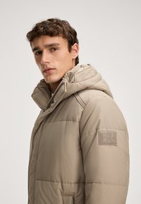 Beżowa kurtka puchowa z gładkiego nylonu, z odpinanym kapturem, pikowaną fakturą i naszywką z logo na lewym rękawie.