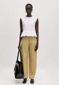 Witte mouwloze top met een afgeronde zoom, beige wijde broek, zwarte schoudertas en zwarte instapschoenen. Accessoires zijn onder andere zwarte zonnebril en oorbellen.