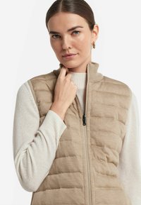 Beige kviltad väst med hög krage, dragkedjestängning och ribbade ärmar i en ljus gräddfärg. Slät textur, minimalistisk design.