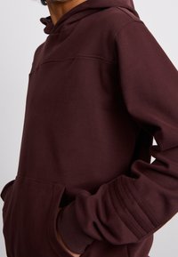 Maroonin huppari, jossa on etutasku, ribattuja yksityiskohtia hihoissa ja kiristysnauha hupussa. Valmistettu pehmeästä kankaasta, jossa on rento leikkaus.