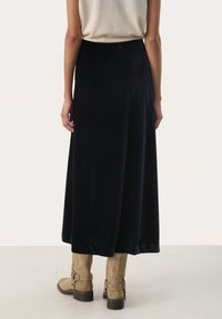 Lunga gonna nera in velluto con una texture liscia, design fluido e vita elastica. Abbinata a stivali alla caviglia color cammello con dettagli a fibbia.