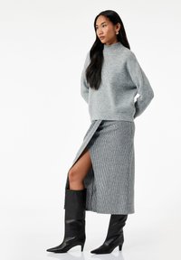 Vrouw in een grijze ribgebreide turtleneck trui en bijpassende rok met een voorkant split, gecombineerd met zwarte kniehoge haklaarzen.