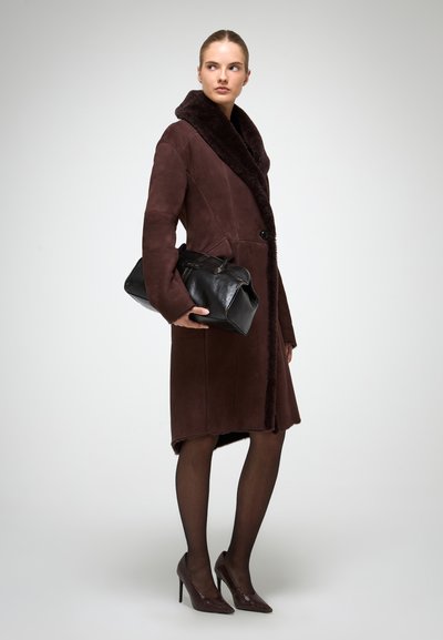 Sieviete valkā tumši brūnu šārlinga mētelis, melnas zeķubikses un strukturētas augstpapēžu kurpes, turot lielu melnu ādas rokassomu, skatoties uz sāniem.