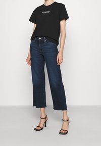 Černé tričko s nápisem "MANKIND", kombinované s tmavě modrými širokými džíny. Modelka má na sobě černé sandály na vysokém podpatku. Jednoduchý a casual design.
