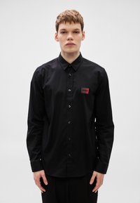 Μαύρο πουκάμισο με κουμπιά από ομαλό ύφασμα, με κλασικό γιακά, μακριά μανίκια και κόκκινο λογότυπο στο αριστερό στήθος.