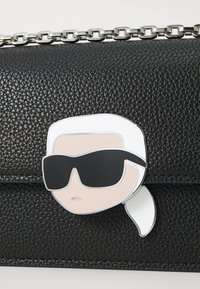 Fekete textúrájú bőrtáska ezüst láncos pánttal, díszítő motívummal, amelyen egy fehér hajú, napszemüveget viselő figura látható.