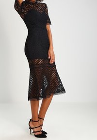 Černé šaty ke kolenům s přiléhavým vrškem a průhlednou krajkovou vrstvou s vzorem. Mají krátké rukávy a rozšířený lem.