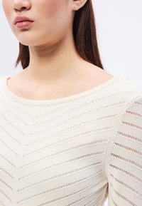 Kremfarget strikket topp med bred hals, som har horisontale striper og åpne vev. Lange ermer med ribbestrikkede mansjetter, myk tekstur.