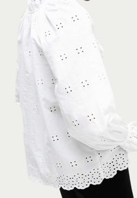 Blouse en coton blanc avec broderie à œillets, manches bouffantes et ourlet festonné. Le tissu a une texture douce et des détails délicats.