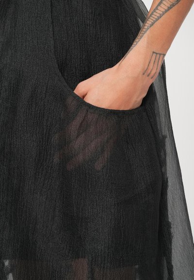 Melnā kleita ar teksturētu, caurspīdīgu audumu. Redzama roka sānu kabatā, demonstrējot apģērba funkcionālo dizainu.