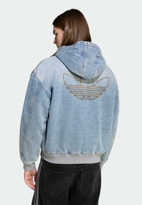 Világoskék farmer kapucnis pulóver kapucnival, arany hímzett logóval a háton, bordázott mandzsettákkal és derékpánttal. Fekete nadrág látható.