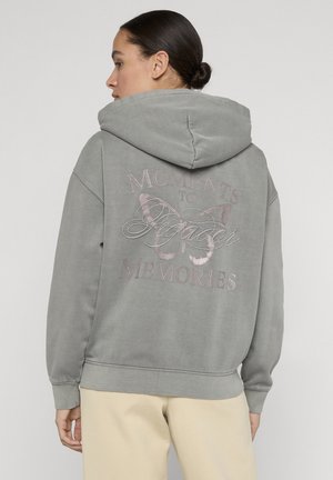 Vrouw met donker haar in een knot, gekleed in een grijze hoodie met een vlinder en de tekst "Moments to Spider Memories" op de achterkant, beige broek.