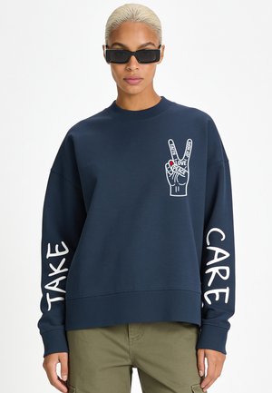 Persoon met kort blond haar en zwarte zonnebril draagt een marineblauwe trui met vredeshand grafiek en de tekst "Take Care" op de mouwen.