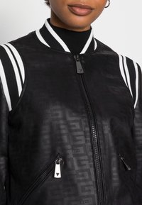 Veste bomber noire avec une finition texturée, col côtelé blanc rayé, et deux poches latérales zippées. Détails en métal argenté sur la fermeture éclair.