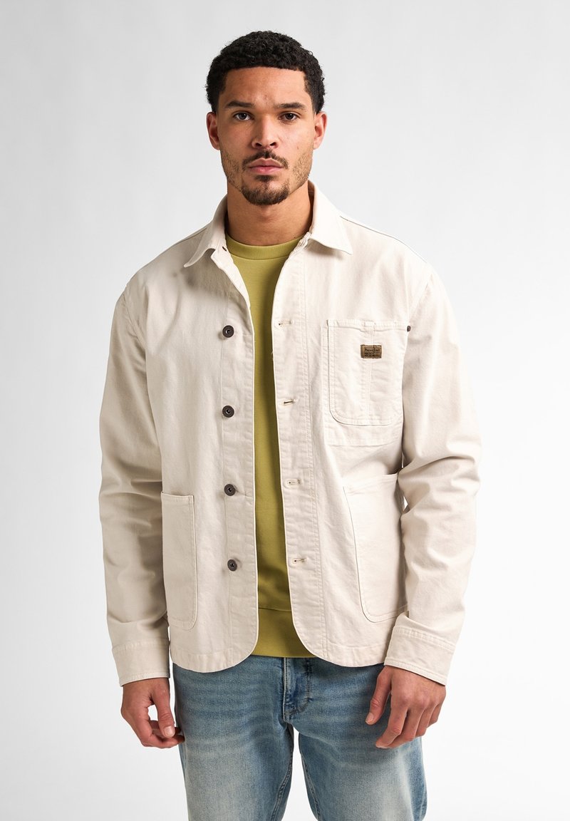 Light beige jacket made of cotton, featuring a button front, two front pockets, and a patch on the chest. Worn over a green shirt, denim jeans.
