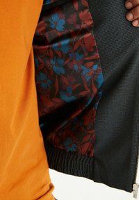 Męska kurtka z czarnym, teksturowanym zewnętrzem oraz żywą, kwiatową podszewką satynową w kolorach czerwonym, niebieskim i czarnym. Elastyczny pas dla dopasowanego kroju.