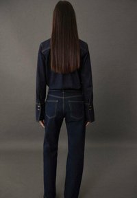 Femme aux longs cheveux raides portant une veste en denim sombre et un jean, se tenant face à un fond gris uni.