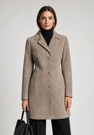 Femme portant un manteau boutonné couleur taupe sur un col roulé noir, tenant un sac à main noir, debout devant un fond clair uni.
