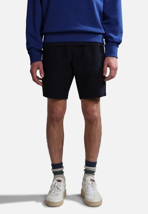 Mand iført sorte shorts, hvide sneakers med slidte hvide snørebånd, stripede sokker og en blå sweatshirt, stående mod en hvid baggrund.