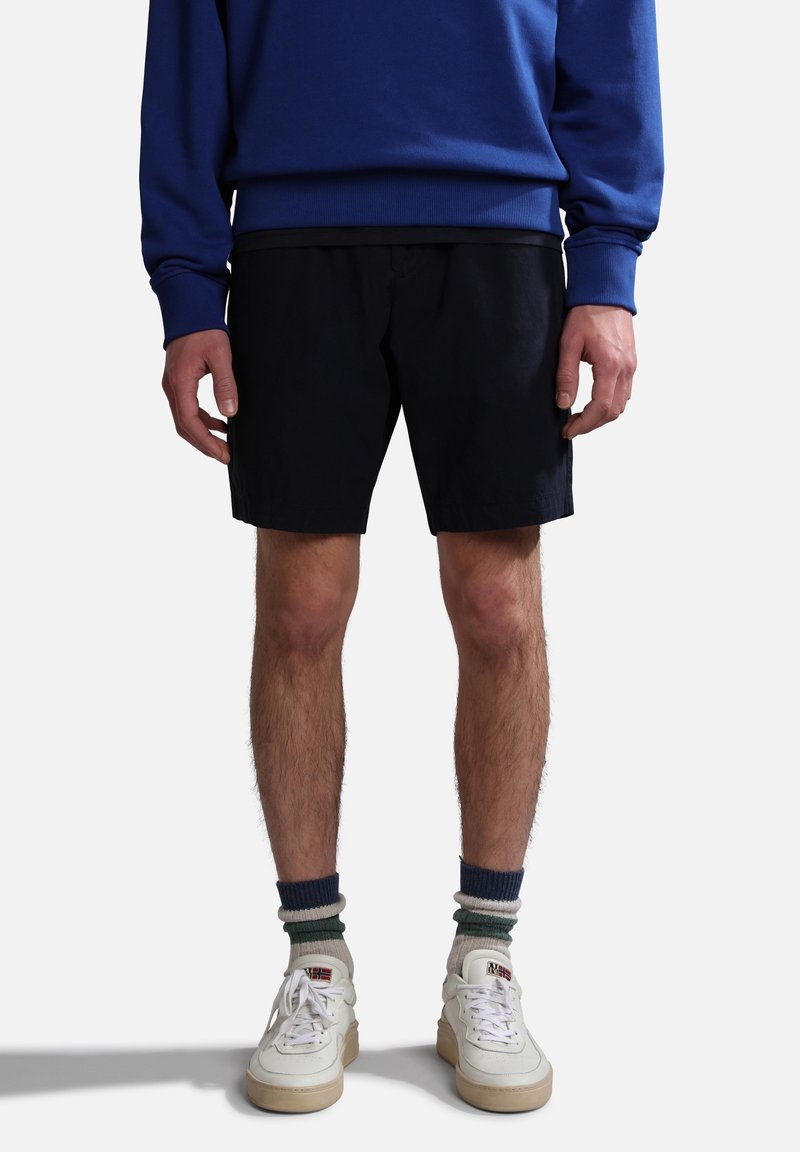 Homme portant un short noir, des baskets blanches avec des lacets blancs usés, des chaussettes rayées et un sweat bleu, debout sur un fond blanc.