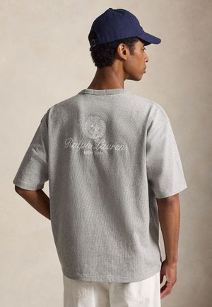 Άνδρας που φορά γκρι μπλουζάκι Ralph Lauren New York και σκούφο σκούρου μπλε, κοιτάζοντας πλάγια με ουδέτερο φόντο.
