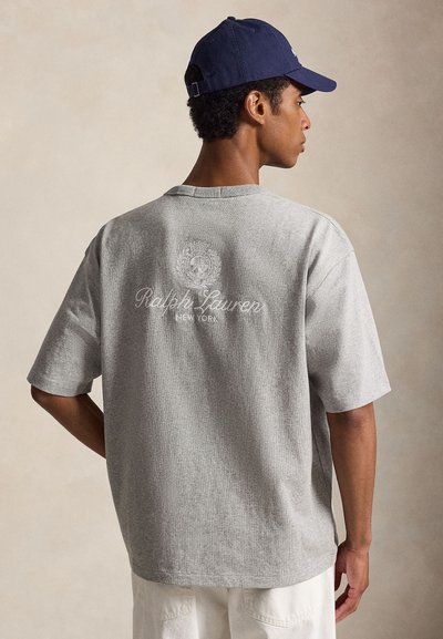Άνδρας που φορά γκρι μπλουζάκι Ralph Lauren New York και σκούφο σκούρου μπλε, κοιτάζοντας πλάγια με ουδέτερο φόντο.