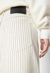 Personne portant un pantalon beige clair à fines rayures verticales noires et une étiquette de marque en cuir noir au-dessus de la poche arrière, associé à un pull crème.