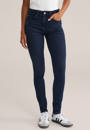 Sötétkék, magas derekú, skinny farmernadrág, denim anyagból készült. Finom kopásnyomokkal és klasszikus öt zsebes kialakítással rendelkezik. Fehér és szürke sportcipőkkel viselve.