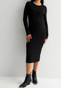 Robe midi noire à manches longues avec un détail de taille nouée, en tissu lisse. Associée à des bottines noires à talon petit hauteur.