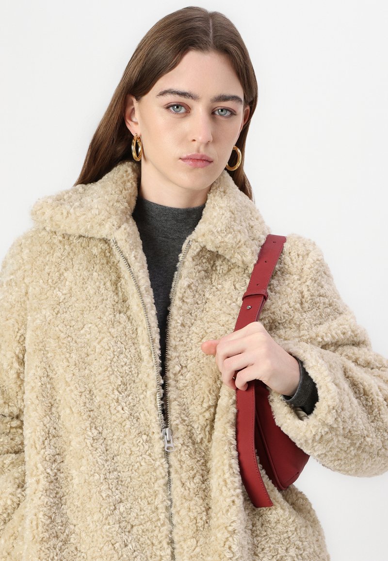 Beige nepbontjas met een rits sluiting en grote kraag. Het model houdt een bordeauxrode schoudertas vast. Grijze coltrui eronder.