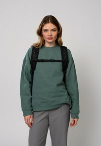 Oversized, leśnozielona bluza z miękkiego materiału, posiadająca okrągły dekolt. Noszona z szarymi spodniami i czarnym plecakiem z regulowanymi paskami.