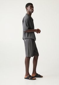 Kortærmet grå skjorte med knapper og grå shorts, med en glat tekstur og afslappet pasform, parret med sorte sandaler med to stropper.
